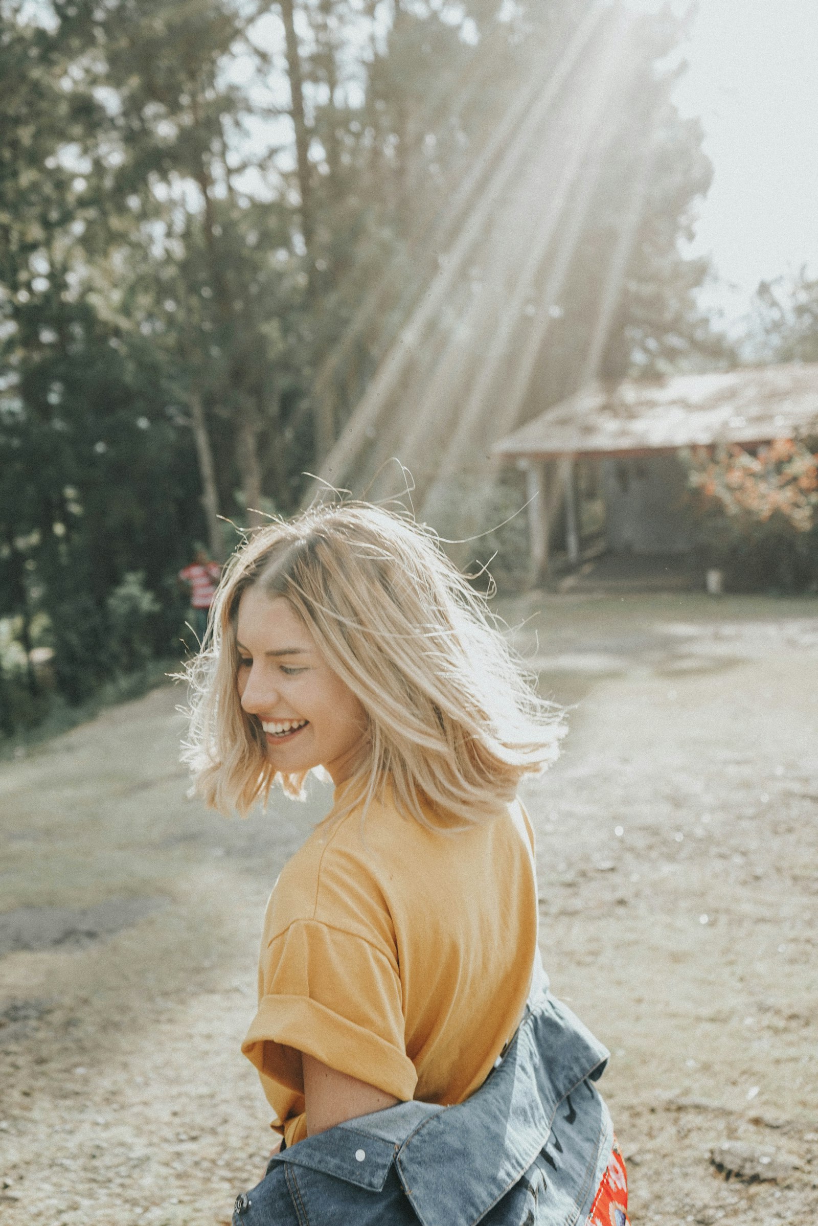 A smiling woman in a yellow shirt outdoors in bright sunlight with trees behind her
