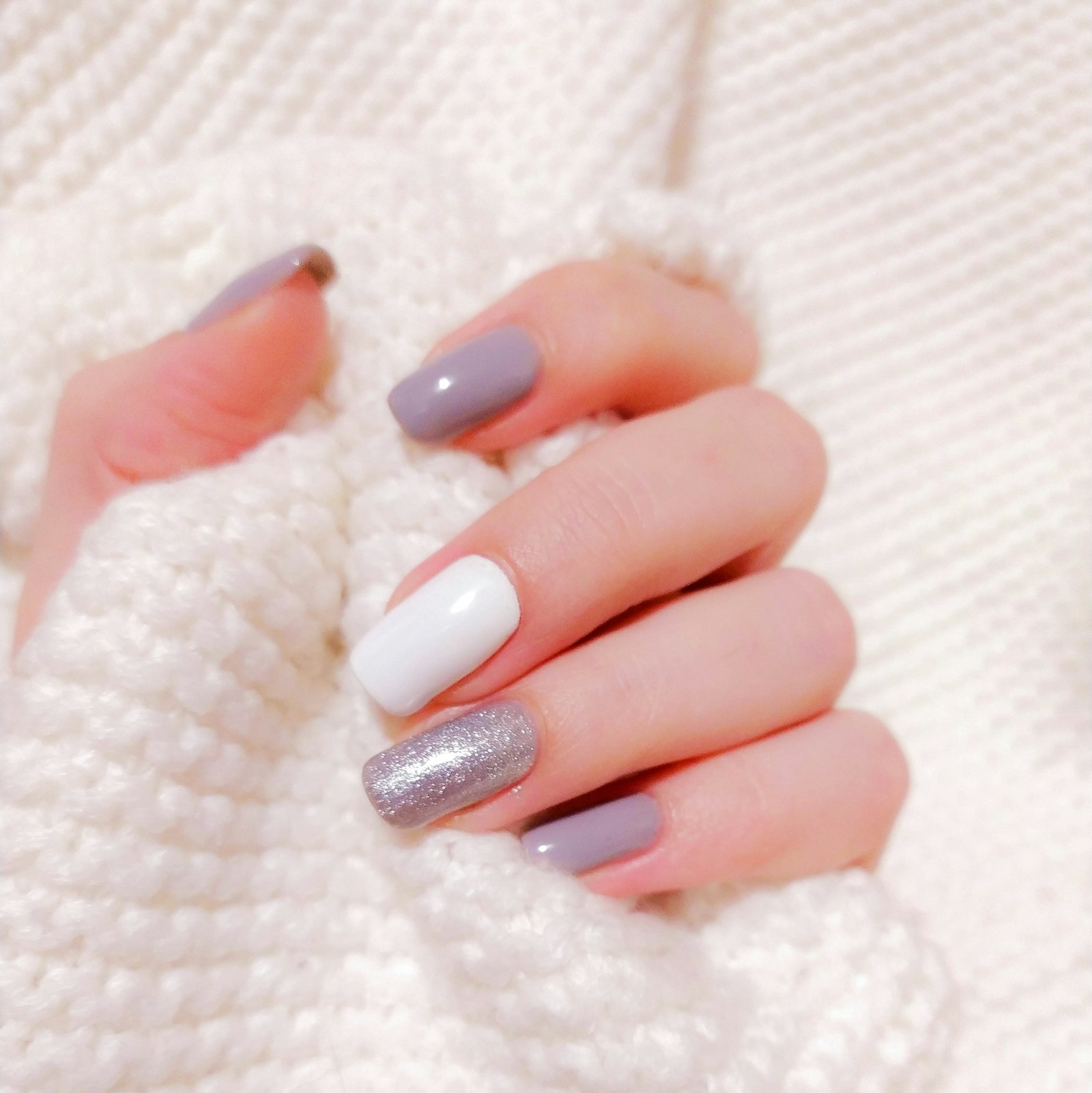 A hand with mixed pastel lavender, white and glitter nails resting on a white knit sweater