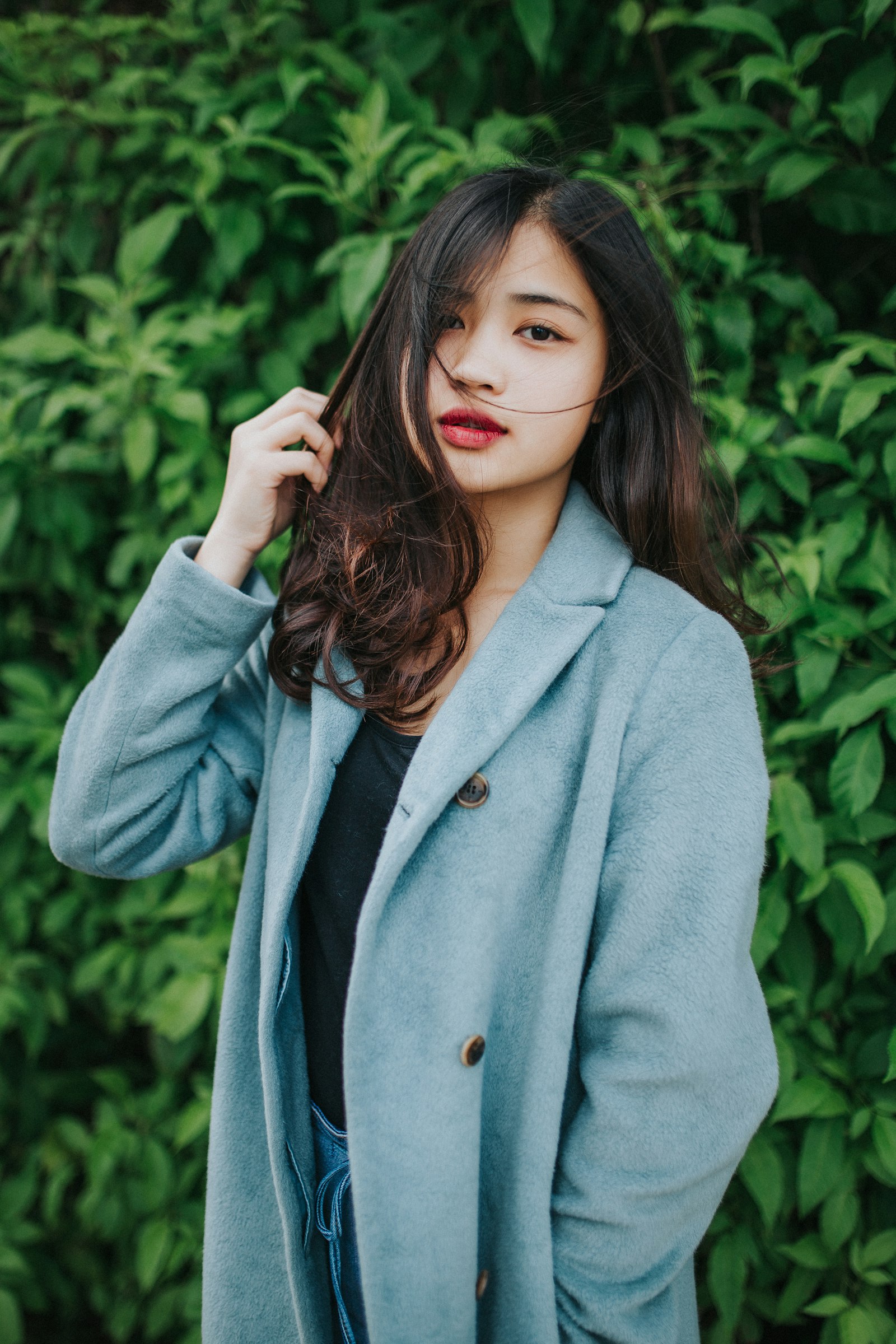 A young woman in a pale blue coat standing against a green hedge